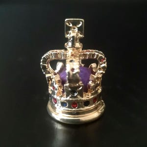 Puede incluir: Un dije de corona en miniatura de tono dorado con un inserto central de color púrpura similar al terciopelo. El marco metálico tiene detalles de pequeñas piedras rojas y azules incrustadas en la base y los arcos, rematado con una cruz.