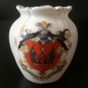 May include: A small white porcelain vase with a fluted rim and gold trim. The front features a colorful heraldic crest with a red shield, black wings, and a gold star. Below the crest, a banner reads "NUNQUAM NON PARATUS" and "MOFFAT".