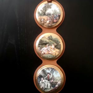 May include: A vertical wall hanging featuring three circular porcelain plaques set in a tan leather strap with a brass buckle. Each plaque displays a colorful pastoral scene with figures in period clothing, framed by a thin gold-toned metal rim.