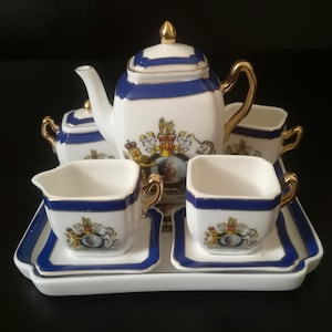 Puede incluir: Un juego de té de porcelana blanca que incluye una tetera cuadrada, un azucarero, una lechera, dos tazas con platillos y una bandeja rectangular. Cada pieza está decorada con un borde azul, detalles dorados y un emblema de escudo real.