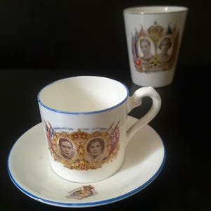 Op de afbeelding: Een wit porseleinen kop en schotel met een blauwe rand, voorzien van portretten van koning George VI en koningin Elizabeth, de tekst Coronation May 12 1937 en een kroonembleem. Een bijpassende witte porseleinen beker staat op de achtergrond.