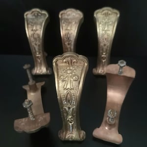 May include: A set of five brass-toned metal drawer pulls featuring an ornate, embossed floral and geometric pattern on the front surface. Each pull has a curved, elongated shape with two threaded metal posts and nuts on the back for mounting.