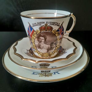 Op de afbeelding: Een wit porseleinen kop en schotel set met gouden rand. De kop is voorzien van een sepia-kleurig portret van koningin Elizabeth II met kinderen, geflankeerd door Union Jack vlaggen en de tekst H.M. QUEEN ELIZABETH II CROWNED JUNE 2ND 1953.