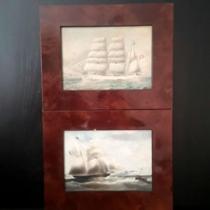 May include: Two stacked rectangular wall art pieces featuring maritime paintings of sailing ships on the ocean. Each painting is encased in a polished, dark reddish-brown faux-wood frame with a glossy finish, displaying white-sailed vessels.