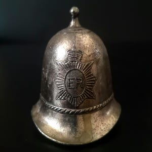 May include: A silver-toned metal bell shaped like a British police custodian helmet. The front features an embossed star-shaped emblem with a crown at the top and the letters ER in the center. A decorative rope-like band encircles the base.