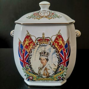 May include: A white ceramic lidded jar featuring a portrait of Queen Elizabeth II, a crown, and two Union Jack flags. The text reads Golden Jubilee, Queen Elizabeth II, and 1952-2002. The lid has a gold-trimmed handle and the ER royal cypher.
