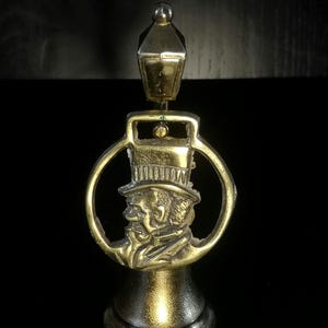 Pode incluir: Um tampão de garrafa de latão com um perfil detalhado de um homem de cartola dentro de uma moldura circular. O tampão inclui um pequeno design de lanterna no topo e uma base decorativa. O design é vintage.