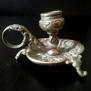 May include: A brass chamberstick candle holder featuring an ornate, embossed floral pattern on the circular base and a matching decorative handle. The candle socket is positioned centrally, and the entire piece has a metallic finish.