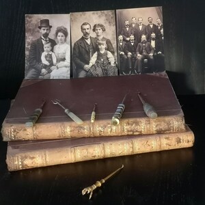 Può includere: Fotografie d'epoca di famiglie e un gruppo di uomini sono esposte sopra due vecchi libri rilegati in pelle. Diversi piccoli strumenti decorativi con manici ornati sono disposti sui libri e uno strumento in ottone a forma di artiglio è sottostante.