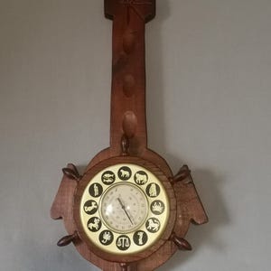 Puede incluir: Un reloj de pared de madera con acabado marrón oscuro. La esfera del reloj es redonda con símbolos del zodiaco alrededor del perímetro. El reloj tiene un tallo largo y decorativo y un diseño circular con pequeñas manijas.