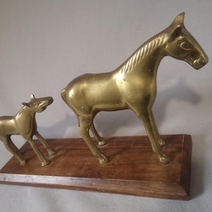 Peut inclure: Deux figurines de chevaux en laiton, un adulte et un poulain, sur une base en bois. Le cheval adulte est debout, tandis que le poulain est dans une pose ludique. Les figurines ont une finition polie et sont de couleur dorée.