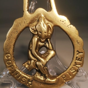 Könnte beinhalten: Ein Vintage-Flaschenöffner aus Messing mit detailliertem Design. Der Öffner zeigt eine sitzende Figur in einem kreisförmigen Rahmen, mit den Worten "Cornish Riskey" am Rand geprägt. Der Öffner hat oben eine Schlaufe zum Aufhängen.