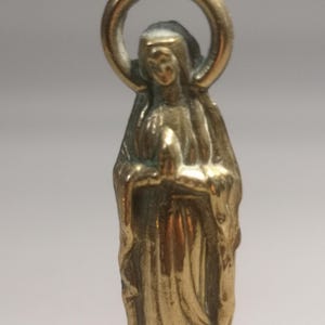 May include: A small, gold-colored statue of a religious figure, likely the Virgin Mary, with hands clasped in prayer. The figure is wearing a draped garment and has a halo above the head. The statue is made of a shiny metal and has a detailed design.