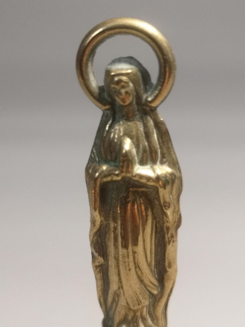 May include: A small, gold-colored statue of a religious figure, likely the Virgin Mary, with hands clasped in prayer. The figure is wearing a draped garment and has a halo above the head. The statue is made of a shiny metal and has a detailed design.