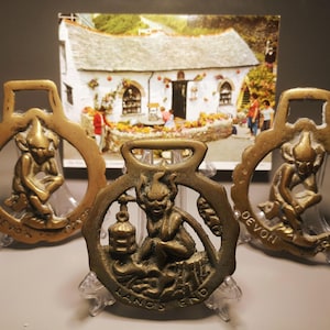 Puede incluir: Tres herrajes de latón antiguos con figuras de duendes. Los herrajes son circulares con un lazo rectangular. Uno dice "LANDS END", los otros "DEVON PIXIE". Una postal de una cabaña está en el fondo.