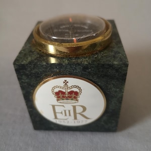 Puede incluir: Un reloj de escritorio de mármol verde con un borde dorado y una parte superior transparente en forma de cúpula. El reloj presenta un emblema circular blanco con una corona y las letras "ER" sobre los números "1952-1977".