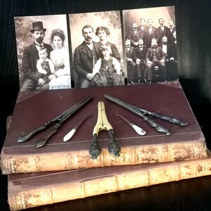 Può includere: Collezione antica con tre ritratti di famiglia vintage e vari strumenti in metallo, tra cui pinze e uno schiaccianoci, disposti sopra due vecchi libri rilegati in pelle. I ritratti sono in bianco e nero.