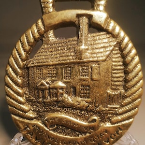 May include: A gold-colored horse brass featuring a detailed relief of a building with a chimney, surrounded by a rope-like border. The text "THE SHIP INN" and "MORWELLHAM QUAY DEVON TAVISTOCK" are inscribed on the brass.