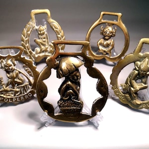May include: Five antique brass horse brasses, each featuring a unique design of a mythical creature. The brasses are in various shapes, with intricate details and the names "POLPERRO" and "DEVON PIXIE" visible.