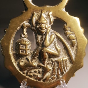 Könnte beinhalten: Ein antikes Pferdegeschirr aus Messing mit einer sitzenden, koboldartigen Figur mit spitzem Hut, die eine Laterne hält. Das Messing hat einen dekorativen Rand und eine Schlaufe zum Aufhängen. Der Kobold sitzt auf einem Baumstumpf.