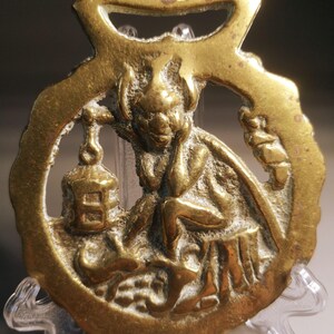 Könnte beinhalten: Ein antikes Pferdegeschirr aus Messing mit einem sitzenden Kobold, der eine Laterne hält. Der Kobold hat Hörner und einen nachdenklichen Ausdruck. Das runde Messing hat oben einen Griff. Es wird auf einem transparenten Ständer präsentiert.