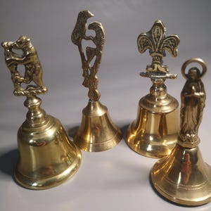 May include: Four antique brass bells with detailed decorative tops. The bells feature a bear, a bird, a fleur-de-lis, and a religious figure. The bells are gold-colored and have a polished finish, showcasing intricate designs and craftsmanship.