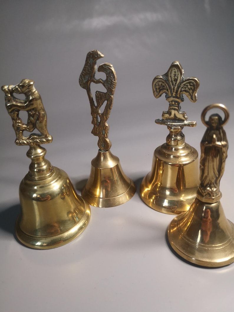 May include: Four antique brass bells with detailed decorative tops. The bells feature a bear, a bird, a fleur-de-lis, and a religious figure. The bells are gold-colored and have a polished finish, showcasing intricate designs and craftsmanship.