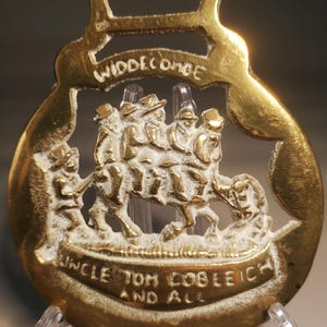 May include: A vintage brass horse brass featuring a relief scene of figures riding a horse-drawn carriage. The words "WIDDECOMBE" and "UNCLE TOM COBLEIGH AND ALL" are inscribed on the brass. The brass is mounted on a clear plastic stand.