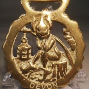 Könnte beinhalten: Ein antikes Pferdegeschirr aus Messing mit einem sitzenden Kobold, der eine Laterne hält. Der Kobold sitzt auf einem Baumstumpf, mit dem Wort "DEVON" unten. Das Messing hat oben eine Schlaufe zum Aufhängen.