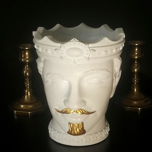 May include: A white ceramic planter shaped like a head wearing a crown. The figure features a winking eye, a gold-painted moustache, and a gold-painted goatee. The crown has a decorative band with circular details. Two brass candlesticks stand behind.