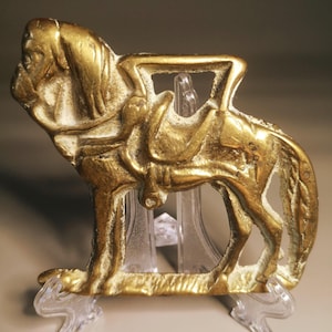 May include: A gold-colored metal sculpture of a horse with a saddle. The horse is in a standing position, with its head turned slightly to the left. The sculpture is detailed, with visible mane, tail, and saddle details. It is displayed on a clear plastic stand.