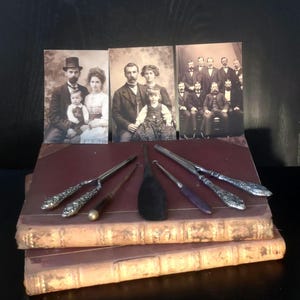May include: A collection of antique items, including vintage photographs and ornate silver utensils, arranged on top of aged, red-bound books. The photographs depict formal portraits of families and groups of people.