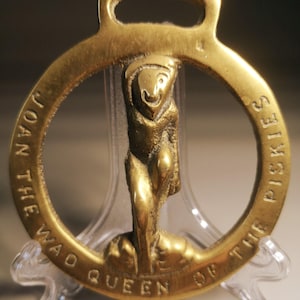 Op de afbeelding: Een vintage messing hanger met een lus aan de bovenkant en een cirkelvormig ontwerp. De hanger heeft een gebeeldhouwde figuur in het midden en de inscriptie "JOAN THE WAO QUEEN OF THE PISKIES" rond de rand. De hanger wordt op een transparante standaard getoond.