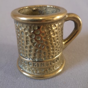 May include: A white ceramic mug with a black handle and the text "But First, Coffee" in a black, handwritten-style font. The mug is on a white surface.
