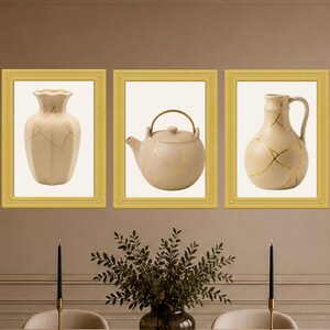May include: Three framed wall art prints featuring off-white ceramic vessels with gold crack detailing. The left print shows a vase, the centre print shows a teapot, and the right print shows a pitcher, all displayed in matching gold frames.