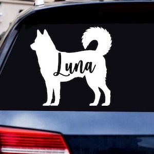 Könnte beinhalten: Ein weißer Vinyl-Aufkleber in Form einer Hunde-Silhouette im Profil mit geringeltem Schwanz, der den Namen Luna in schwarzer Schreibschrift in der Mitte des Hundekörpers zeigt und auf einer dunklen Fahrzeugscheibe angebracht ist.