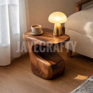 Puede incluir: Una mesa auxiliar de madera con una forma orgánica única. Presenta un acabado marrón cálido y un estante inferior. Una pequeña lámpara con forma de hongo con base dorada y una taza de café sobre un platillo están encima. El texto "JAYECRAFTY" es visible.
