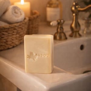 Puede incluir: Una pastilla de jabón cuadrada, blanquecina, con la palabra "SABOU" y un diseño de hoja tallado en la parte delantera. El jabón está sobre el borde de un lavabo blanco, con una vela encendida y toallas enrolladas en el fondo.