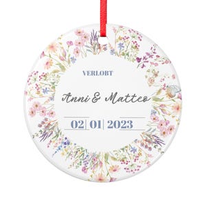 Op de afbeelding: Een witte keramische ornament met een bloemenkrans ontwerp. Het ornament heeft de tekst "VERLOBT", "Anni & Matteo" en de datum "02|01|2023". Een rood lint is bevestigd om op te hangen.