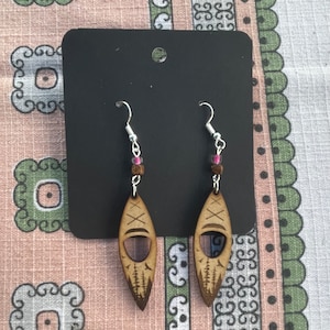 Könnte beinhalten: Holz-Ohrhänger mit Kanu-Design. Die Ohrringe haben eine hellbraune Oberfläche mit geätzten Details, darunter eine Baumreihe und Kreuzmarkierungen. Sie sind an silberfarbenen Haken mit rosa und braunen Perlen aufgehängt.