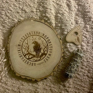 May include: A wooden planchette and a round wooden board with a raven design and the text "YES", "NO", and "FAREWELL". The board has the alphabet and numbers around the edge. A bundle of sage is also present.