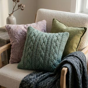 Könnte beinhalten: Ein gemütlicher Sessel mit vier dekorativen Kissen und einer gefalteten Decke. Die Kissen sind in Grün- und Lavendeltönen gehalten, eines davon mit Zopfmuster. Eine graue Decke mit dezentem Muster ist über dem Arm drapiert.