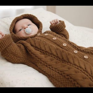 May include: A baby in a brown knitted hooded jumpsuit with wooden buttons and a dummy. The jumpsuit features a cable knit pattern and is lying on a white patterned blanket.