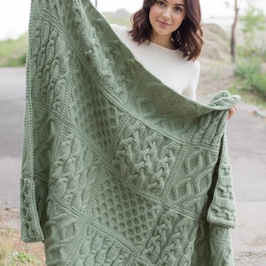 Könnte beinhalten: Eine salbeigrüne Strickdecke mit verschiedenen strukturierten Mustern, darunter Zopfmuster und Wabenmuster. Die Decke ist rechteckig und scheint handgefertigt zu sein, mit einem weichen, gemütlichen Aussehen. Die Decke wird gehalten.