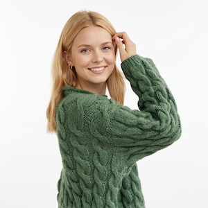 以下が含まれることがあります： ハイネックの緑色のケーブルニットセーター。セーターは、織り込まれたテクスチャードデザインです。モデルは笑顔で、金髪です。背景は白です。