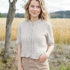 May include: A beige short-sleeved knit cardigan featuring a textured diamond pattern, a vertical cable knit detail along the button placket, a ribbed crew neckline, and ribbed sleeve cuffs. The garment is fastened with small, light-colored buttons.