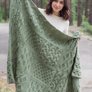 Puede incluir: Una manta tejida de color verde salvia que presenta una compleja disposición de patrones texturizados, incluyendo trenzas verticales, motivos de diamantes y texturas de panal. El tejido rectangular está hecho con hilo grueso y color uniforme.