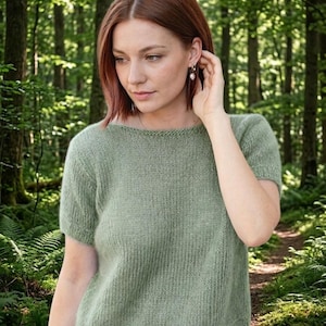 May include: A short-sleeved, sage green knit top. The top has a crew neck and a ribbed texture. The model is wearing the top in a forest setting. The top is made of a soft, comfortable material.