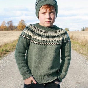 Könnte beinhalten: Ein salbeigrüner Strickpullover und eine passende Mütze mit einer runden Passe, die ein geometrisches Muster in Creme, Dunkelbraun und Schwarz aufweist. Der Pullover hat einen gerippten Rundhalsausschnitt, Bündchen und Saum, die Mütze einen Ripprand.