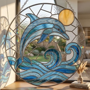 May include: A circular stained glass suncatcher featuring a blue dolphin leaping above stylized blue ocean waves. The design includes a yellow sun in the upper right corner, all encased in a metal frame with a silver chain for hanging.